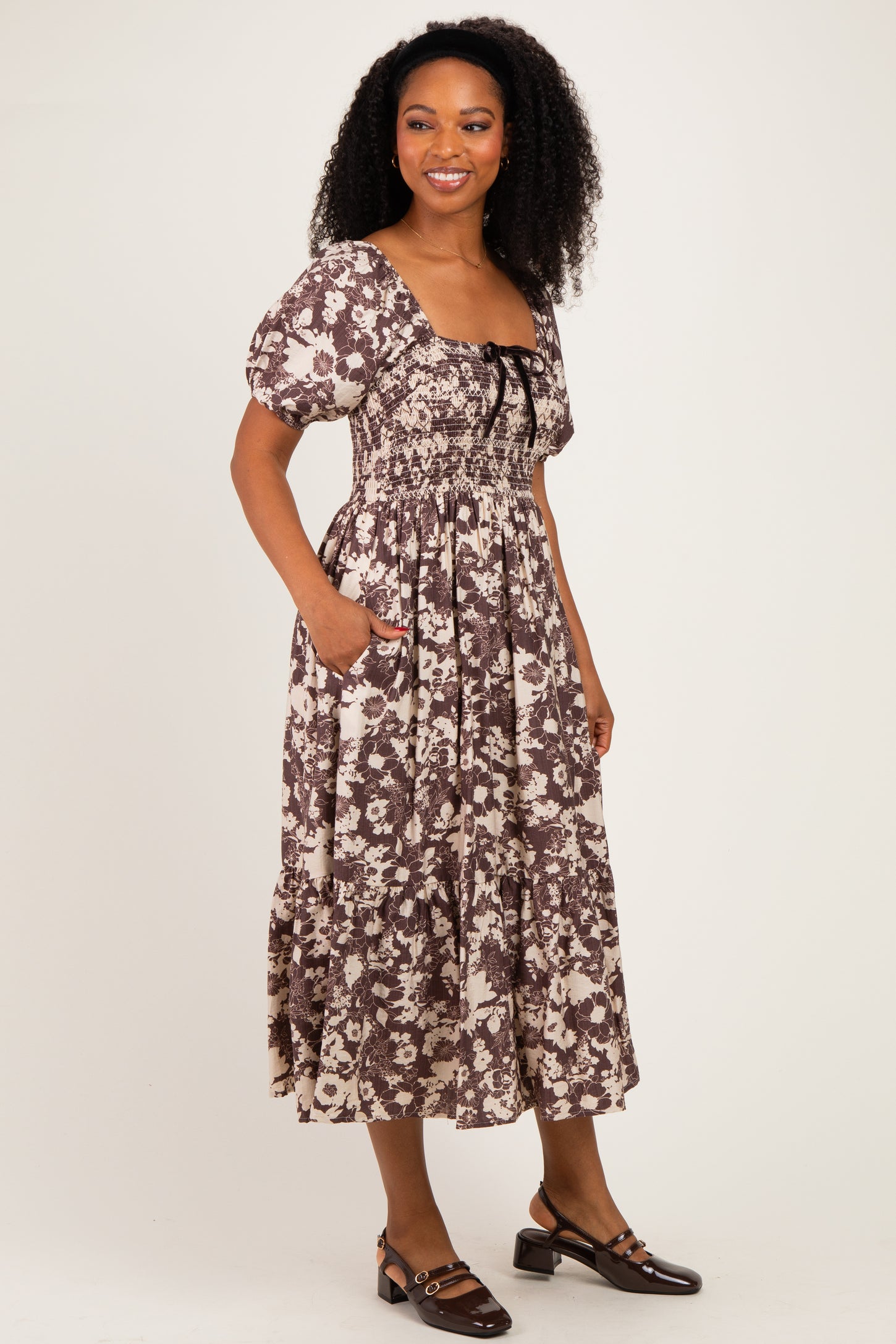 woman wearing a brown floral midi dress with puff sleeves and a square neckline, standing with hands in pockets