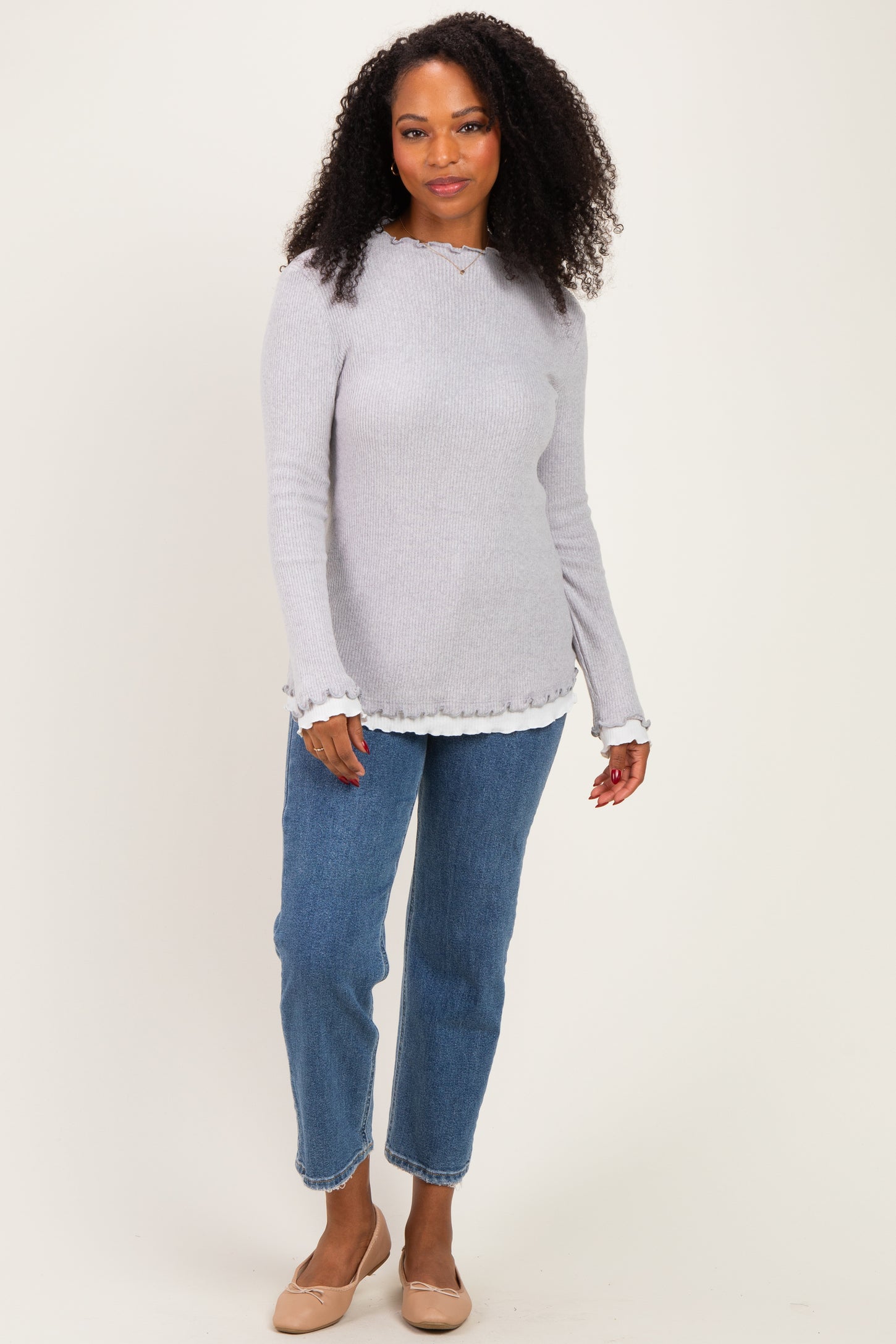 woman wearing a light gray ribbed long sleeve sweater with ruffle details and blue high-waisted jeans
