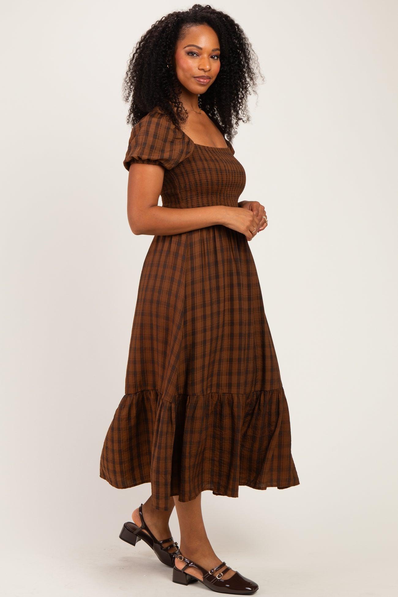 woman wearing a brown plaid midi dress with puff sleeves and a ruffled hem, standing against a neutral background