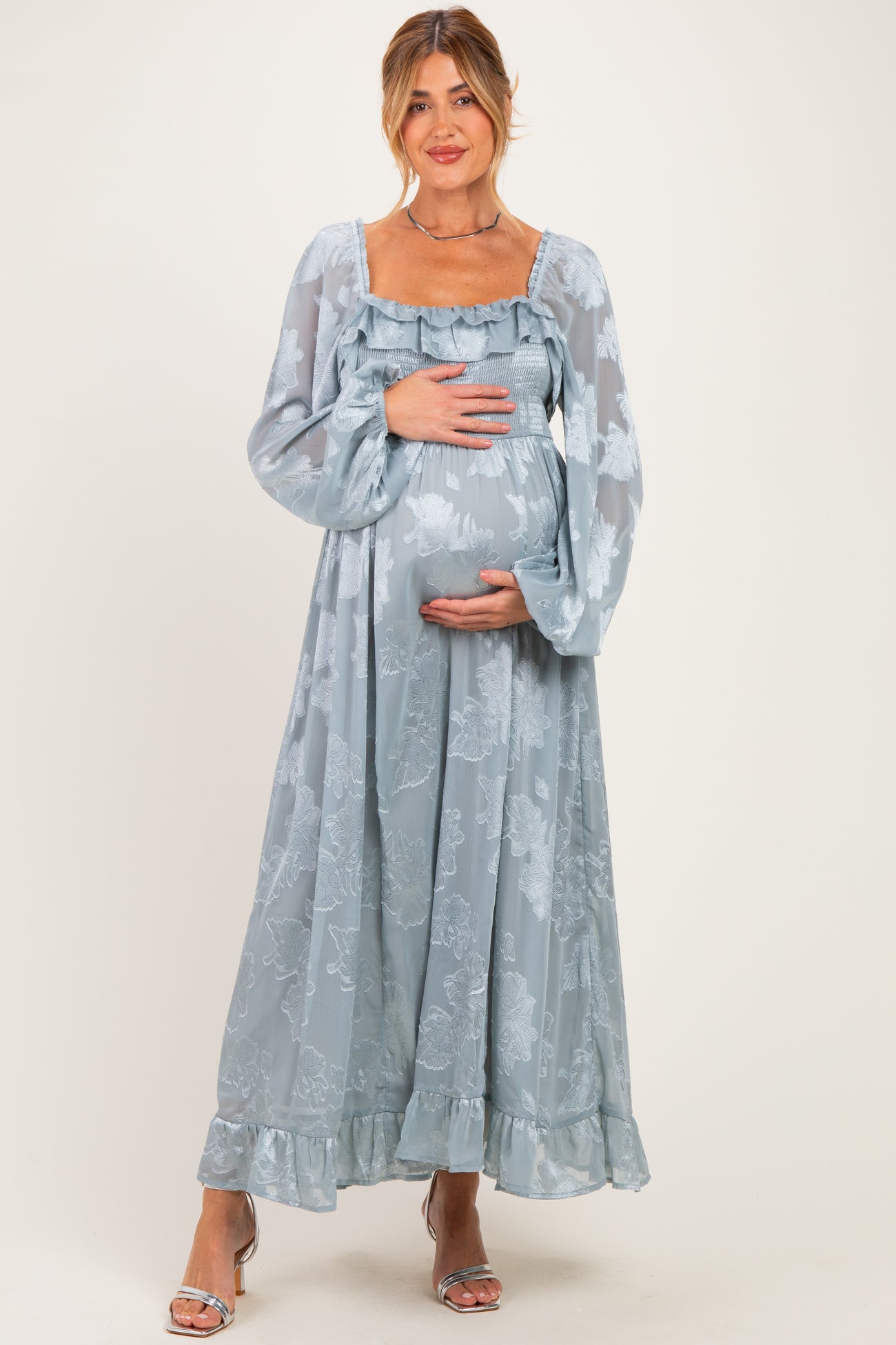 woman wearing a long light blue maternity dress with floral patterns and a smocked bodice, holding her baby bump