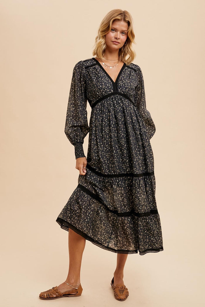 woman wearing a black floral tiered midi dress with long sleeves and a deep v-neckline, standing against a neutral background