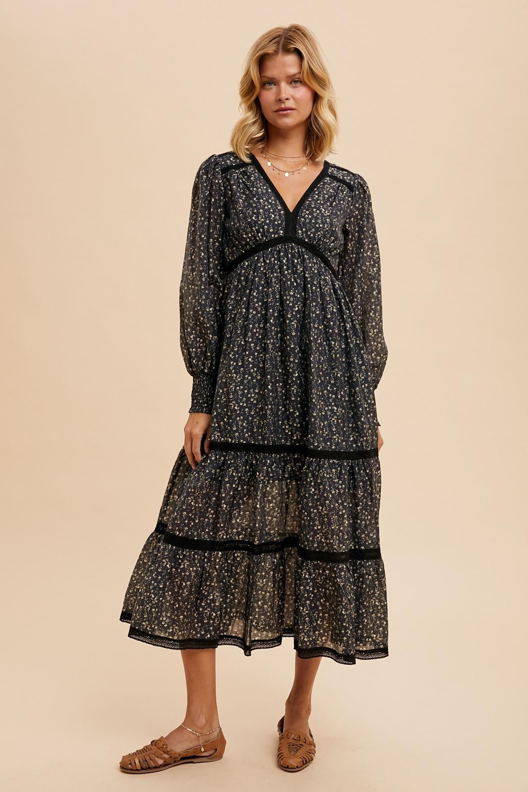 woman wearing a black floral tiered maxi dress with a v-neckline and long sleeves, standing on a neutral background