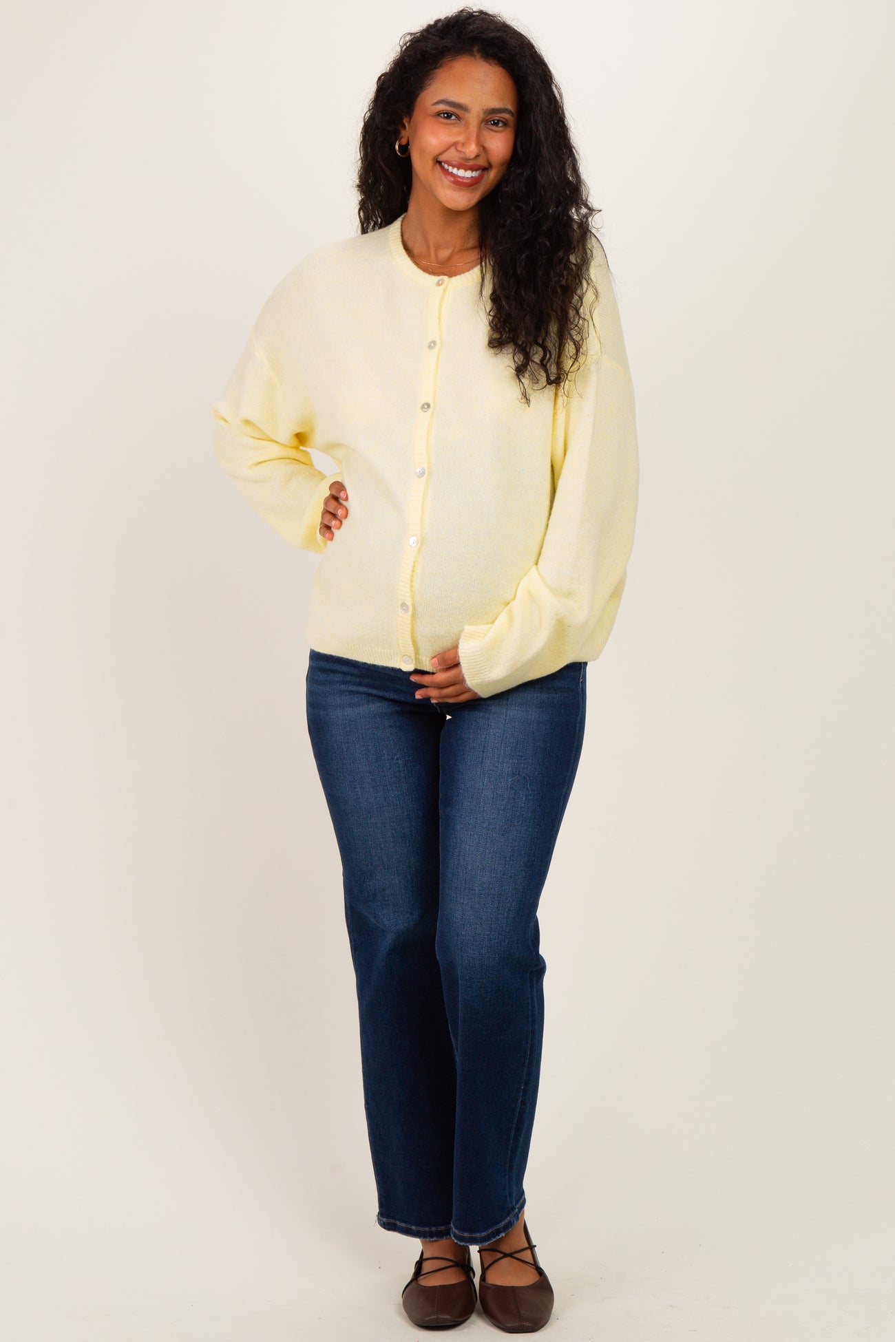 woman wearing a yellow maternity cardigan and blue jeans, gently holding her baby bump while smiling