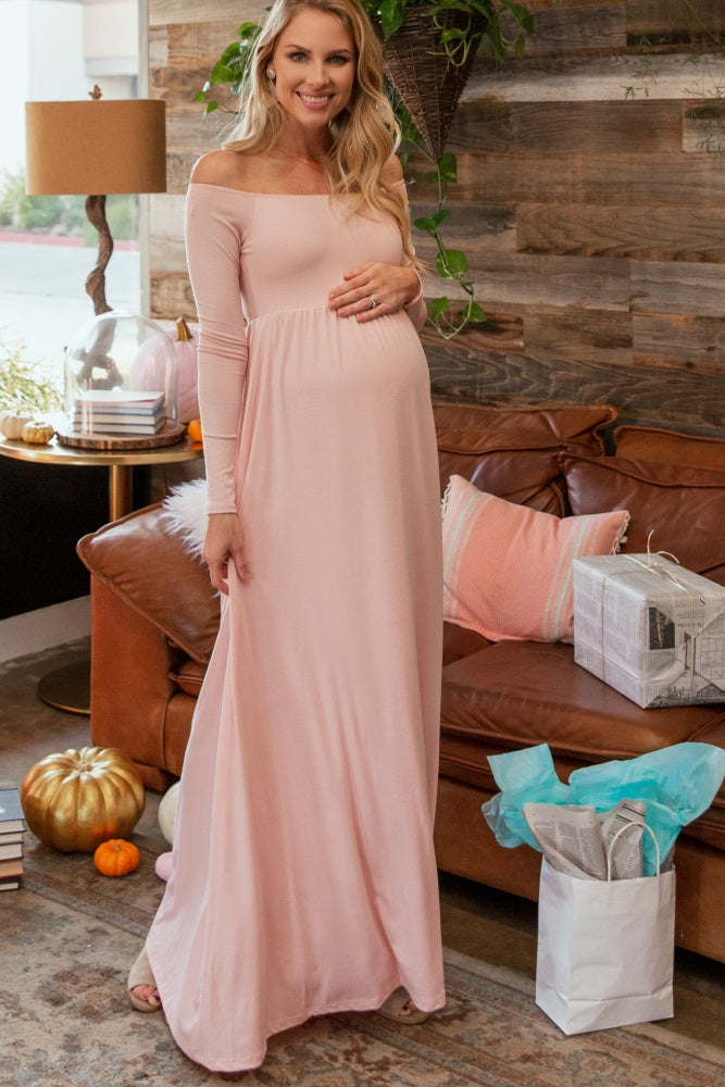 woman wearing a light pink off-shoulder maternity maxi dress and holding her baby bump while standing in a cozy living room