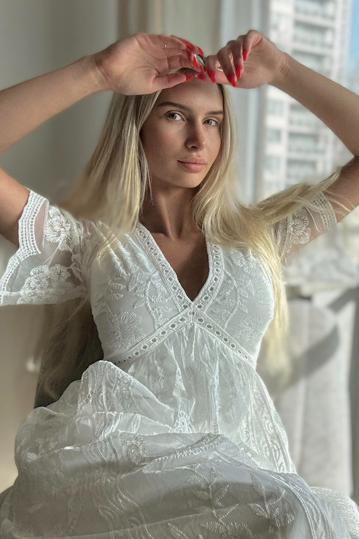 woman wearing a white lace maxi dress with short sleeves, sitting and posing gracefully by a window