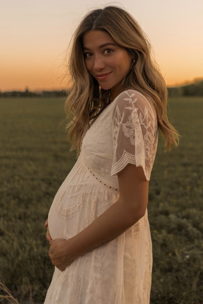 woman wearing a cream maternity dress with lace details, showing her baby bump against a sunset backdrop