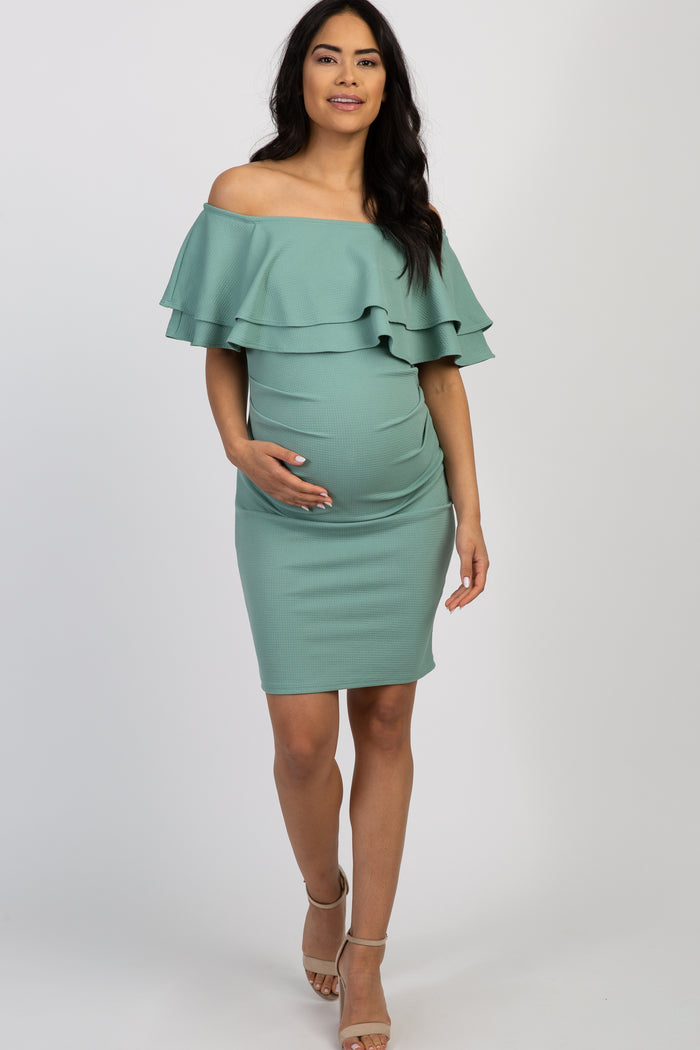 woman wearing a sage green off-the-shoulder ruffled maternity dress, holding her baby bump