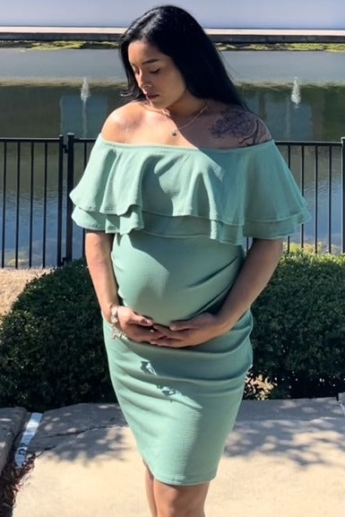 woman wearing a light green off-shoulder maternity dress with ruffle details and holding her baby bump