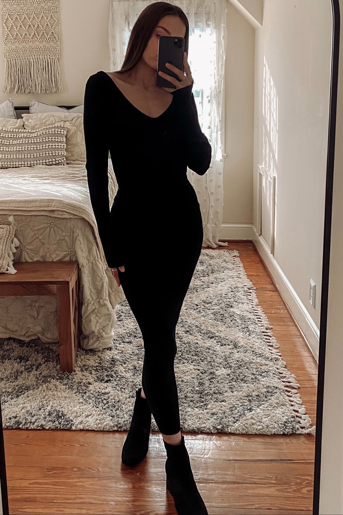woman wearing a black long sleeve fitted dress and ankle boots, posing in front of a mirror in a cozy bedroom setting