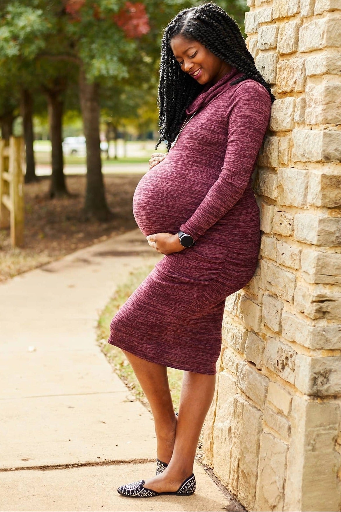 woman wearing a burgundy maternity dress with ruched details and a visible baby bump, leaning against a stone wall