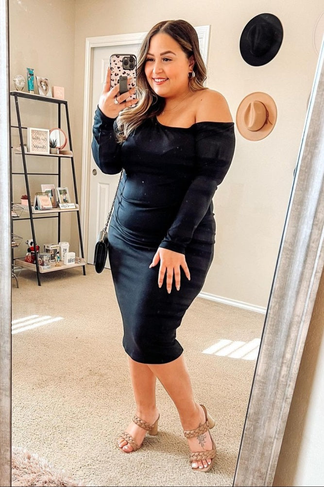 woman wearing a black off-the-shoulder fitted dress and standing in front of a mirror, smiling, with strappy heels