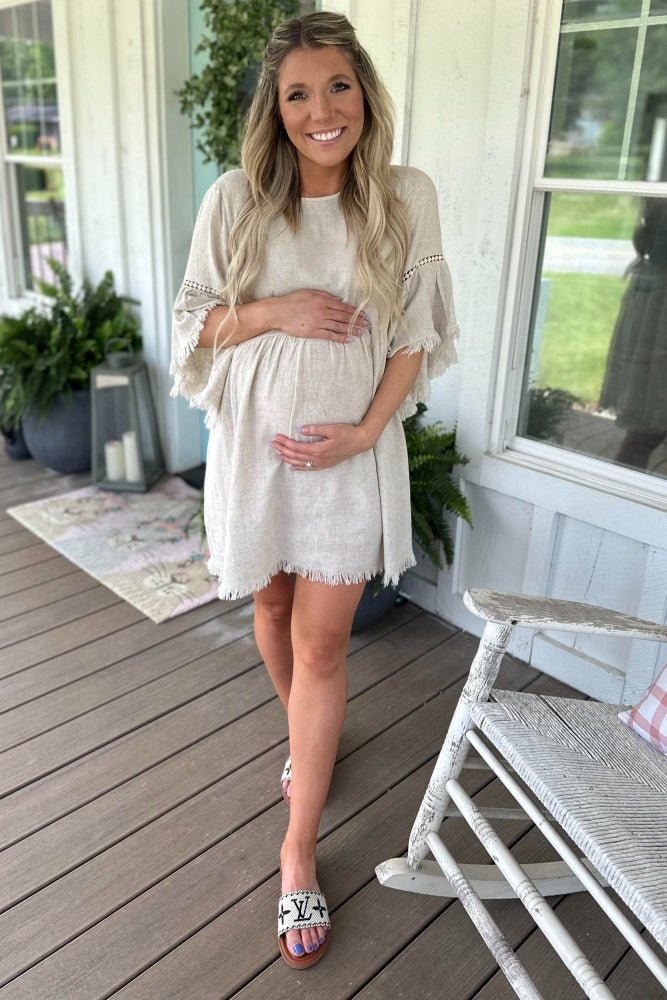 woman wearing a beige maternity dress with fringe details and holding her baby bump while standing on a porch