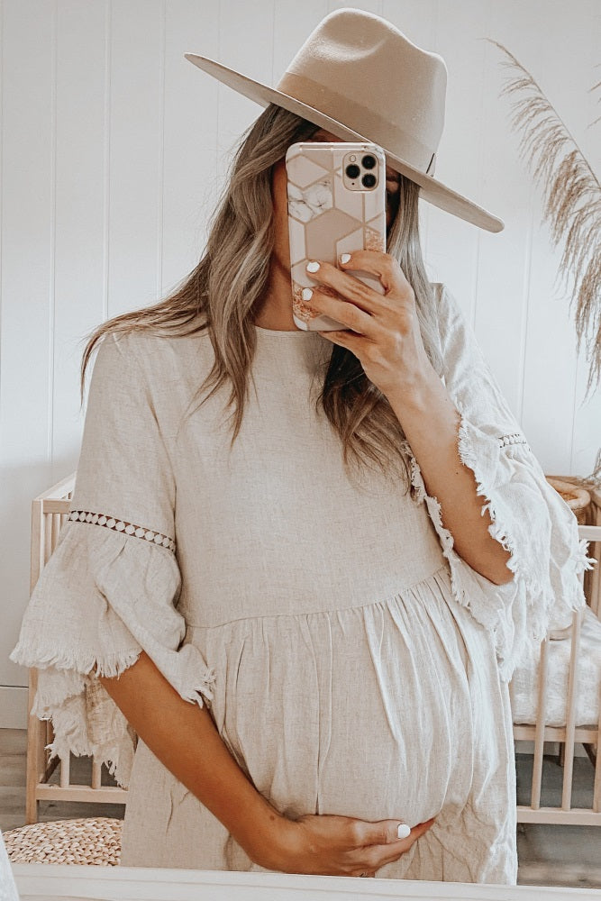 woman wearing a beige maternity dress with ruffled sleeves and holding her baby bump
