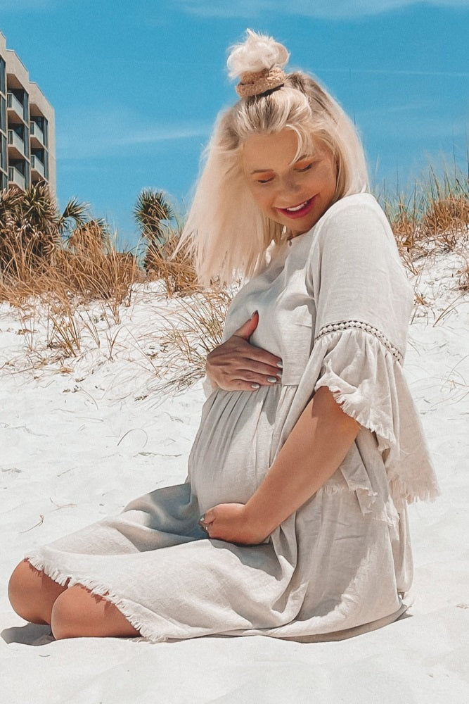 woman wearing a light beige maternity dress with ruffle sleeves and a visible baby bump, seated on the beach