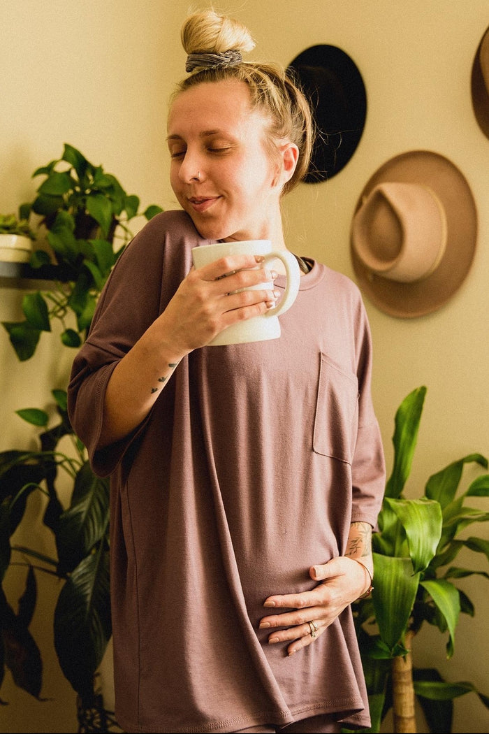 woman wearing a dusty rose oversized maternity top and holding a mug while gently cradling her baby bump