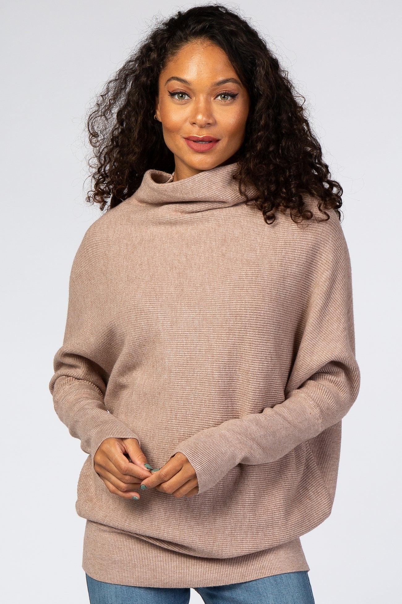 woman wearing a light brown oversized sweater with a cowl neck and long sleeves, paired with blue jeans