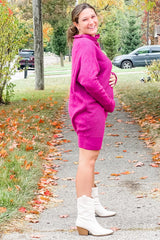 woman wearing a fuchsia maternity sweater dress and holding her baby bump, standing on a leaf-covered path