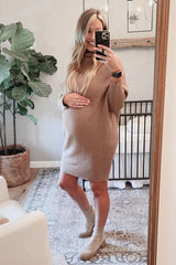 woman wearing a beige maternity sweater dress and holding her baby bump while standing in a room with plants