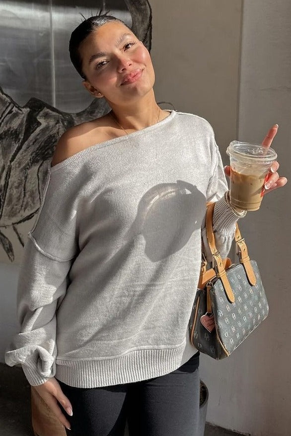 woman wearing a light gray off-shoulder sweater with rolled sleeves, holding an iced coffee and a designer handbag