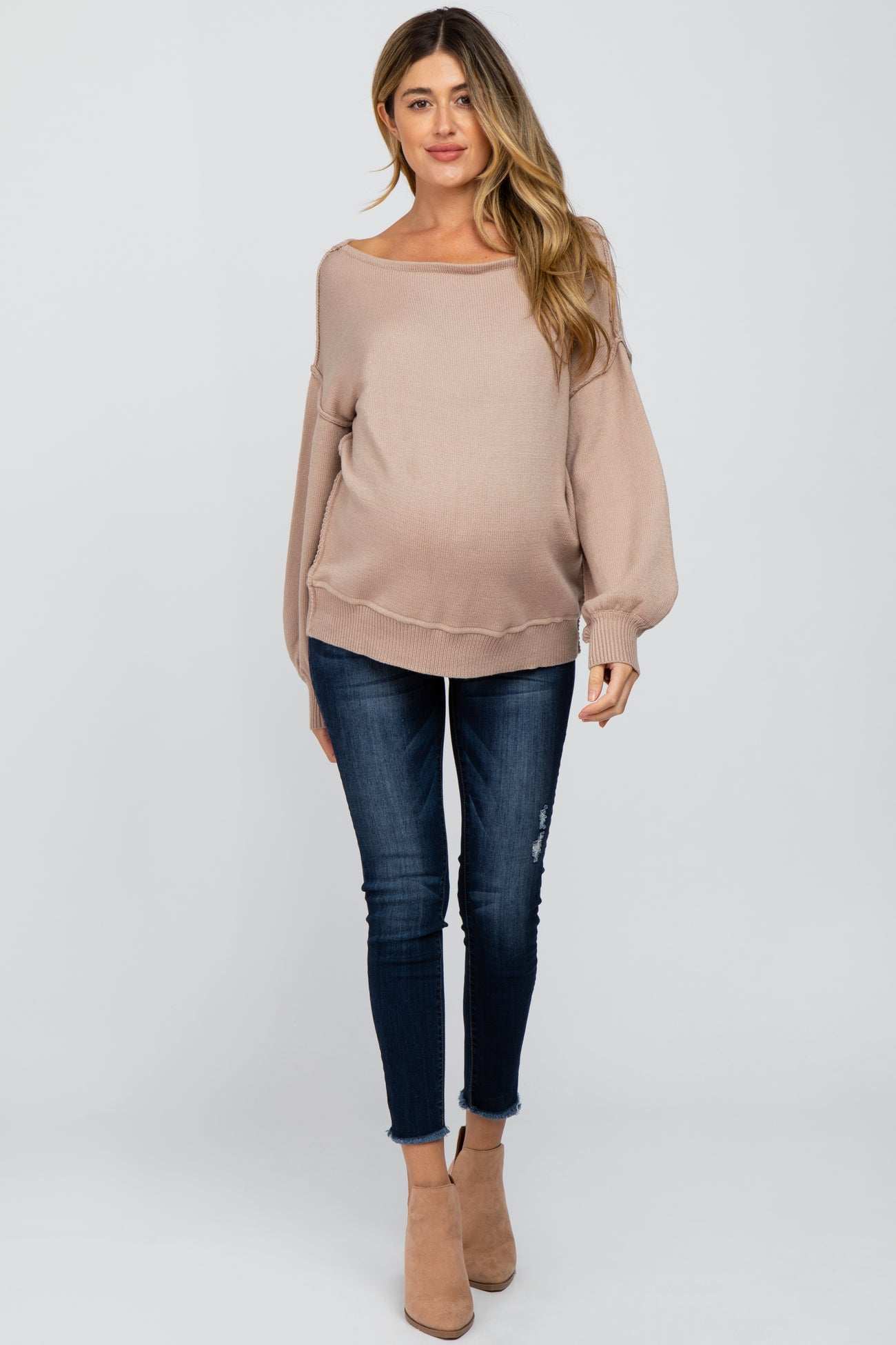 woman wearing a light beige oversized maternity sweater paired with dark blue skinny jeans and beige ankle boots, showcasing a baby bump