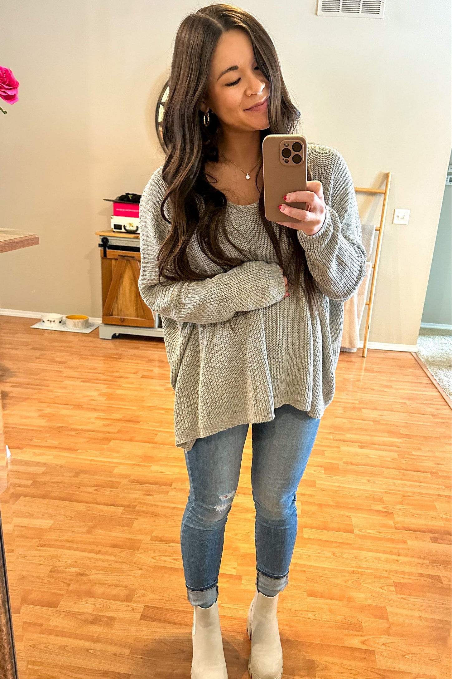 woman wearing a light gray oversized maternity sweater and skinny jeans, smiling while holding her baby bump in a mirror selfie