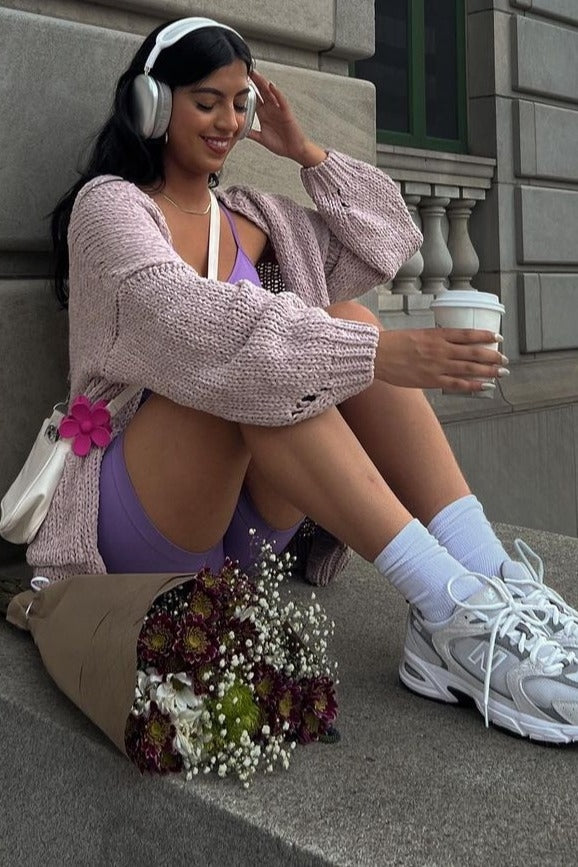woman sitting on steps wearing a lavender bodysuit, oversized pink cardigan, sneakers, and holding a coffee cup with flowers nearby