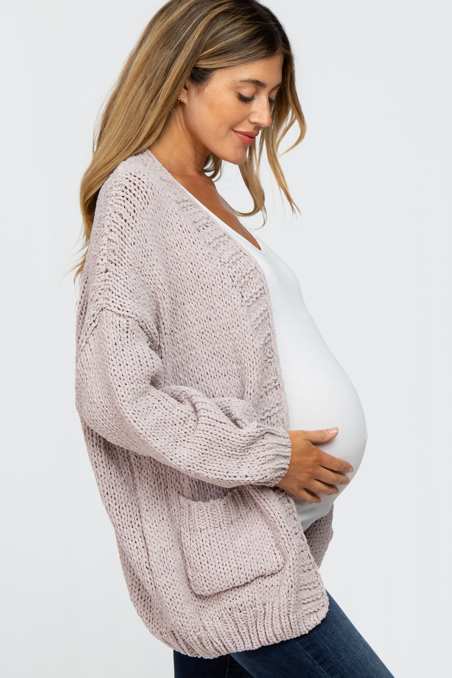 woman wearing a light pink maternity cardigan, holding her baby bump, paired with a white top and dark jeans