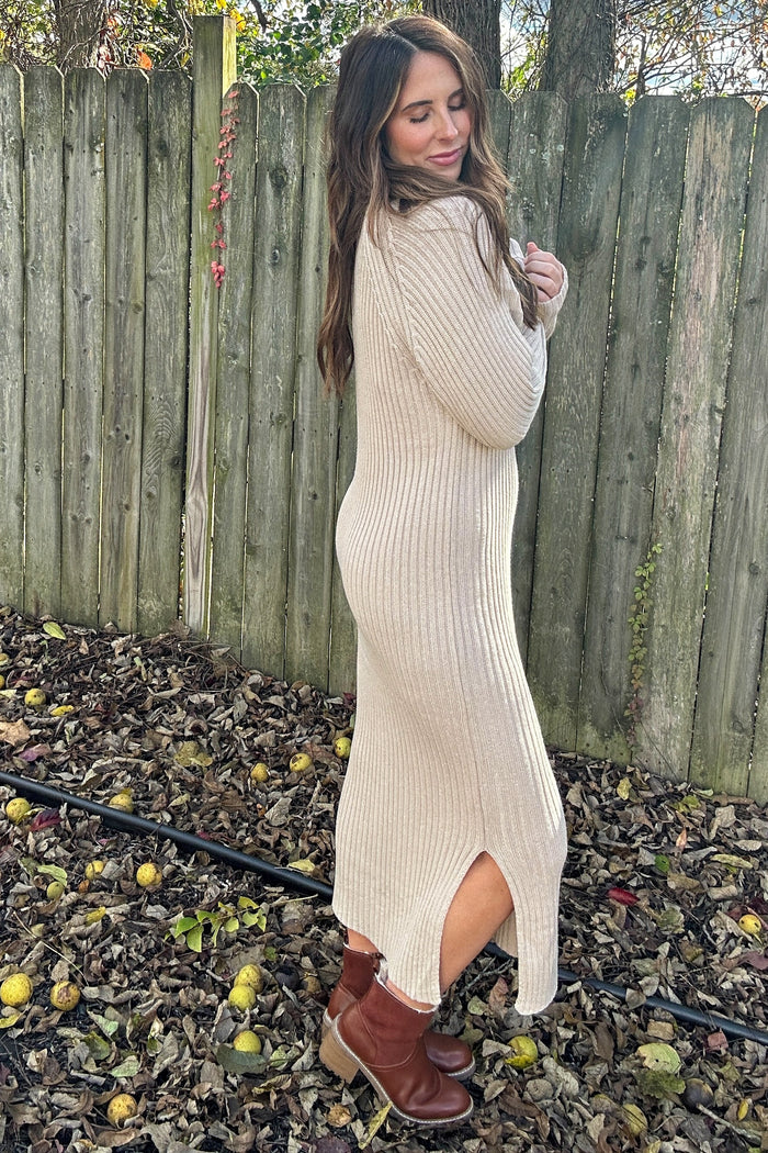 woman wearing a beige ribbed sweater dress with side slits and brown ankle boots, standing outdoors among fallen leaves