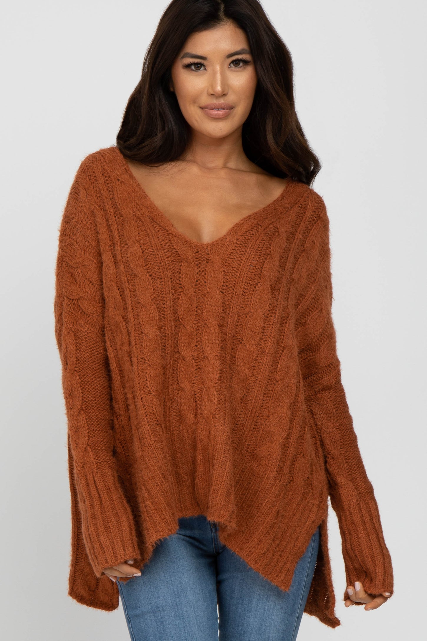 woman wearing an oversized rust cable knit sweater with a deep v-neckline and high-low hem, styled with blue jeans