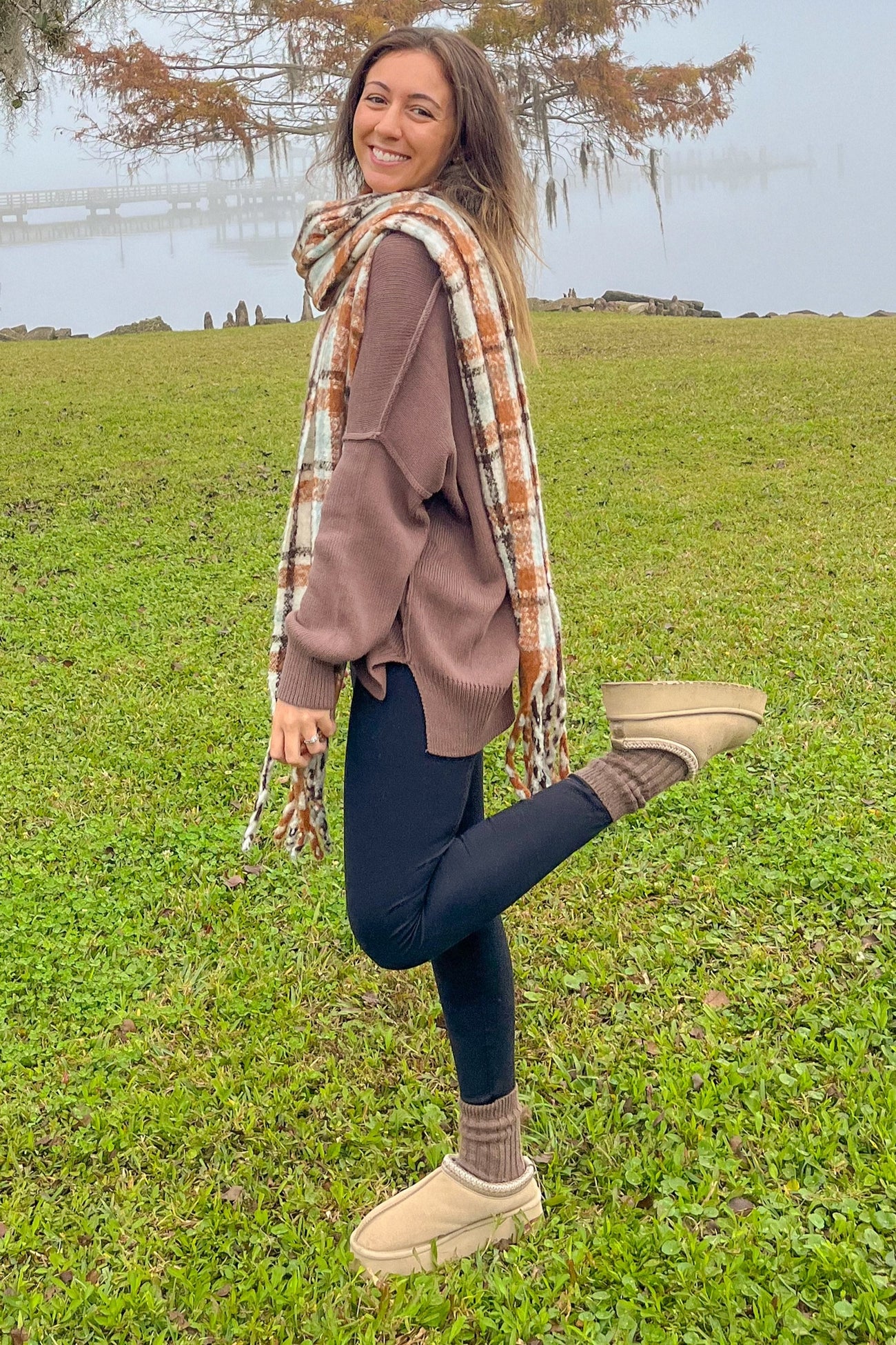 woman wearing a brown oversized sweater, black leggings, and tan ankle boots, wrapped in a plaid scarf, posing outdoors