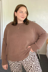 woman wearing a brown oversized knit sweater and black and white patterned pants, smiling slightly with hands on hips