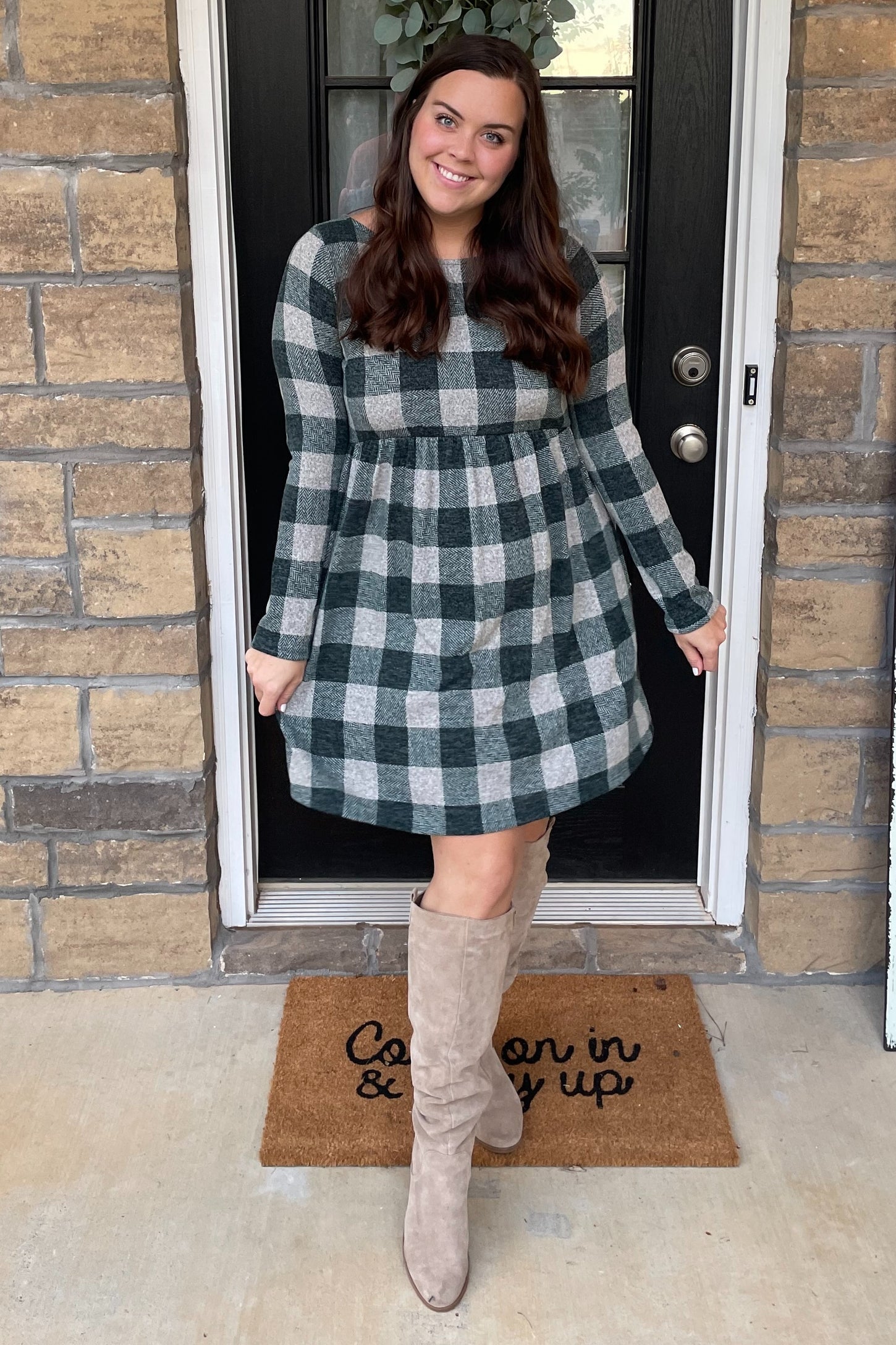 woman wearing a green and gray plaid maternity dress with a baby bump, standing on a porch next to a welcome mat