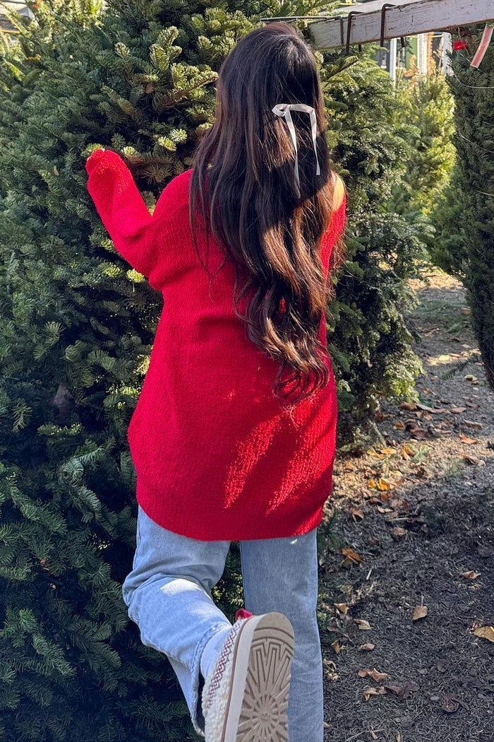 woman wearing a slouchy red sweater and light blue jeans standing next to a tree