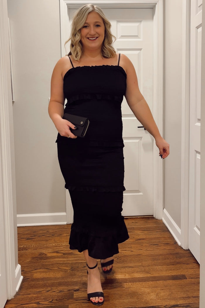 woman wearing a black ruffled midi dress with spaghetti straps and holding a small clutch bag