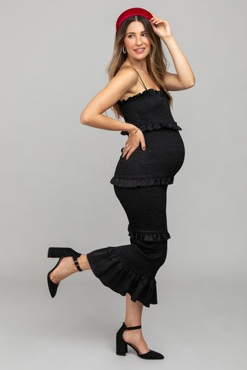 woman wearing a black maternity dress with ruffle details, proudly showing her baby bump and posing with one leg lifted