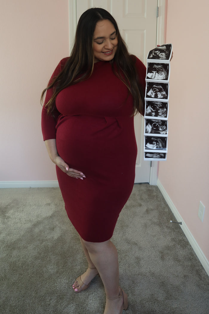 woman wearing a burgundy three-quarter sleeve maternity dress and holding ultrasound photos with a visible baby bump