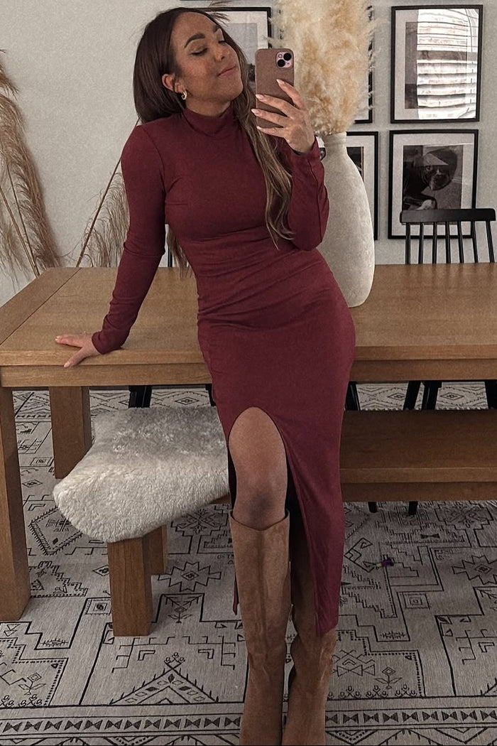 woman wearing a burgundy long sleeve fitted dress with a side slit and knee-high tan boots, posing in front of a mirror