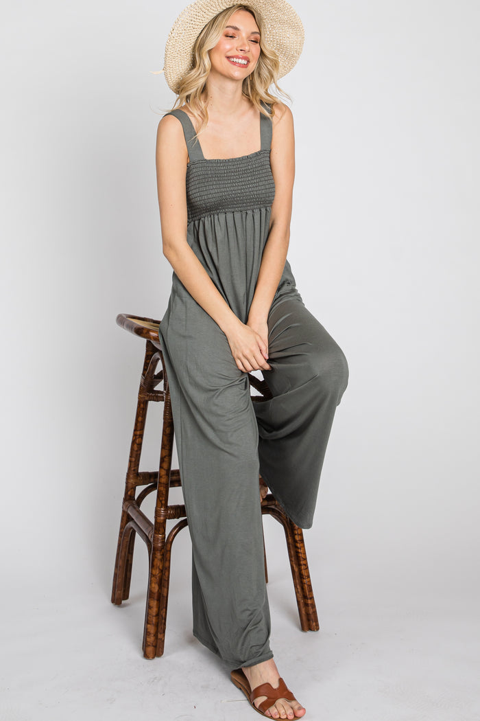 woman wearing a green open-back jumpsuit with a smocked bodice and relaxed fit, seated on a stool