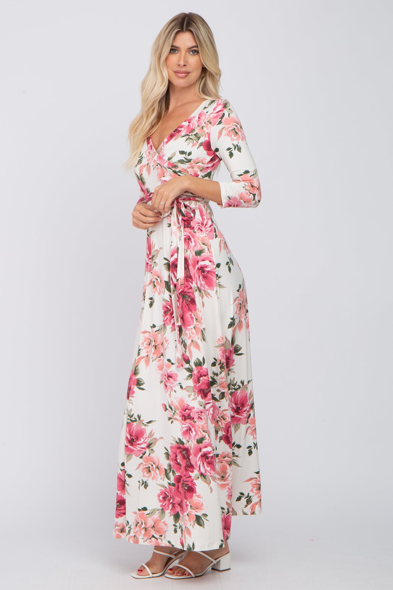 woman wearing a white floral maxi dress with pink roses and long sleeves, standing and holding the dress ties