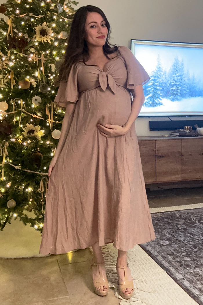woman wearing a blush pink maternity maxi dress with a tie detail at the bust and holding her baby bump