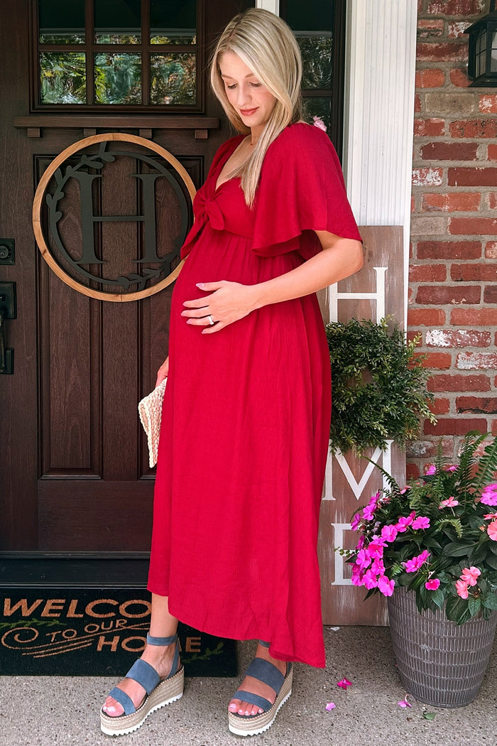 woman wearing a red maternity maxi dress with a bow detail, holding her baby bump while standing at a front door