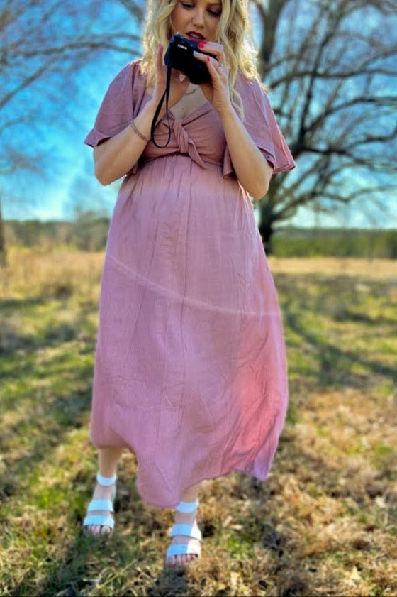 woman wearing a light pink maternity dress with short sleeves and a tie at the waist, holding a camera and showing her baby bump