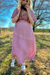 woman wearing a light pink maternity dress with short sleeves and a tie at the waist, holding a camera and showing her baby bump