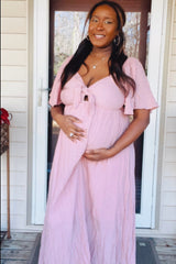 woman wearing a light pink maternity dress with short flutter sleeves and a tie front, gently cradling her baby bump