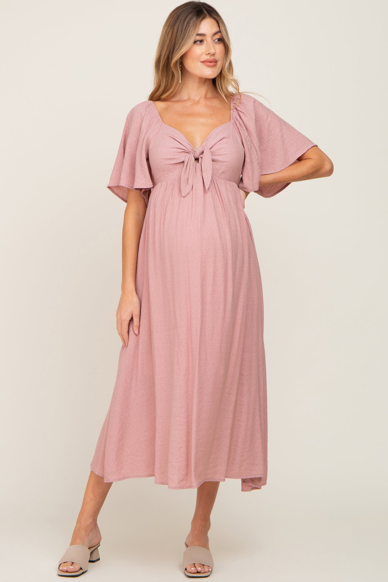 woman wearing a pink maternity maxi dress with a tied front detail and flutter sleeves, showcasing her baby bump