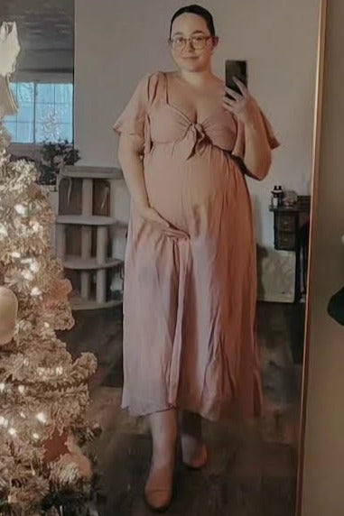 woman wearing a pink maternity dress with a tie front and pockets, showing her baby bump, standing beside a decorated Christmas tree