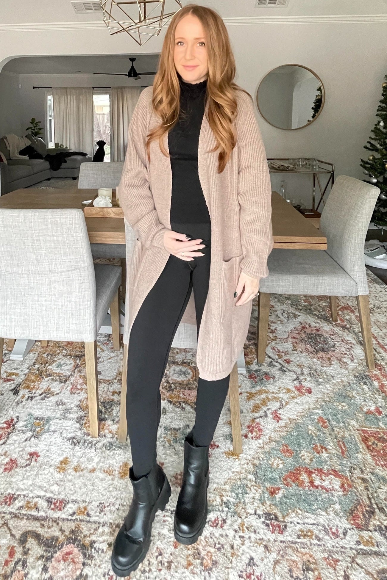 woman wearing a black long sleeve maternity top and leggings, with a beige cardigan, holding her baby bump