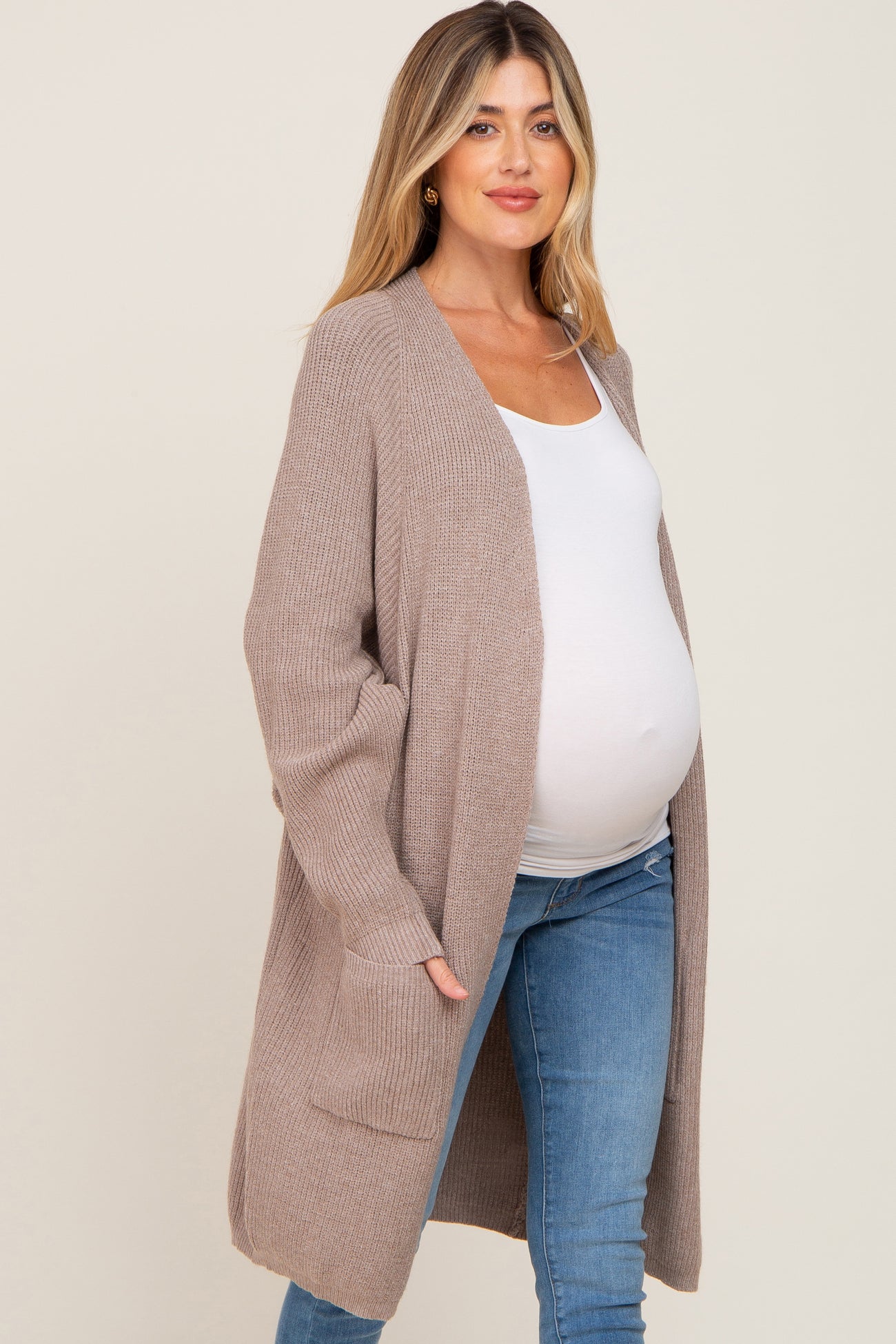 woman wearing a beige long sleeve maternity cardigan over a white top and showing her baby bump while smiling