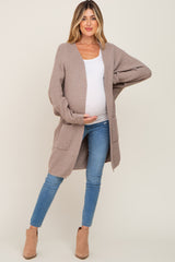 woman wearing a tan open-front maternity cardigan, white top, and blue jeans, holding her baby bump
