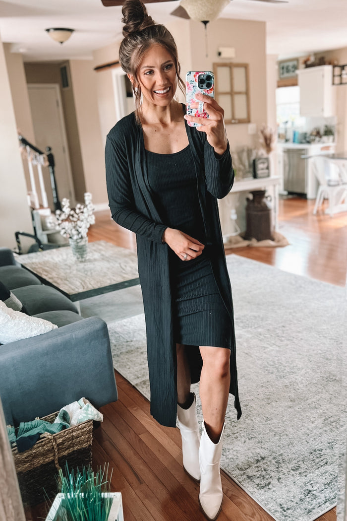 woman wearing a black ribbed dress with a long black cardigan and white ankle boots, standing in a cozy living room